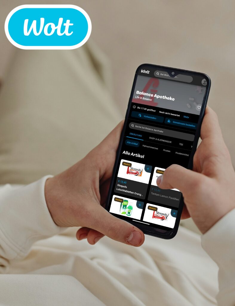 Hände mit Smartphone auf der Website von Wolt im Shop der Balance Apotheke mit hellem Pullover und Jogginghose